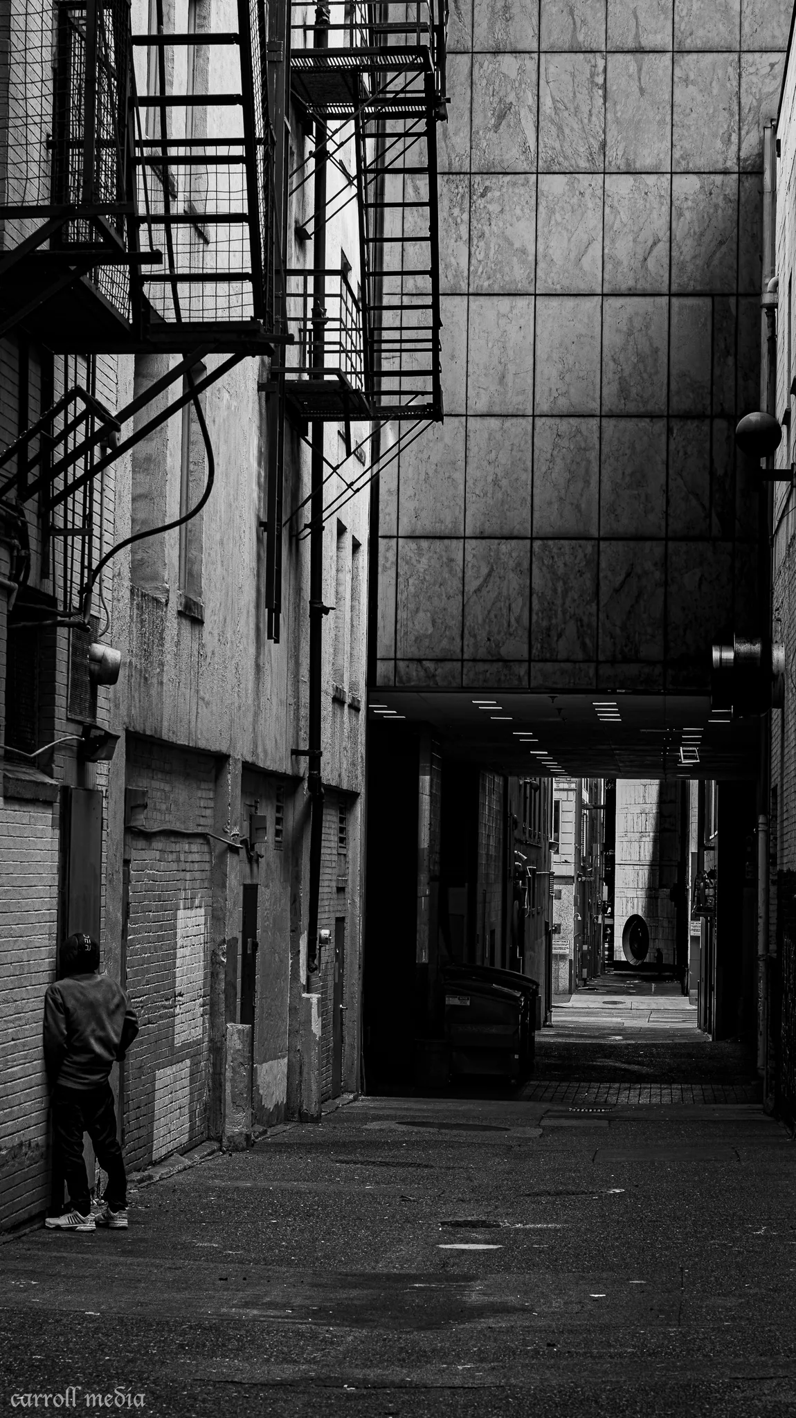 Monochrome alley with fire escape and a lone figure against brick.