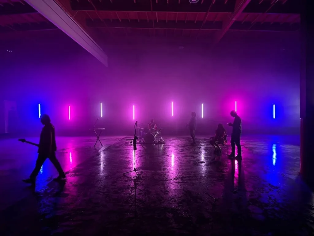 Silhouetted band and gear in a hazy studio with vertical purple and blue LED tubes.