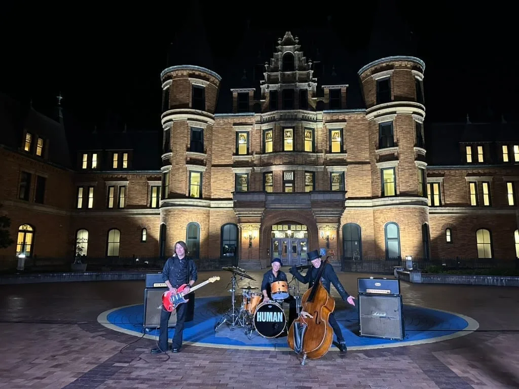 Three-piece band performing at night on a courtyard in front of a lit historic brick building.